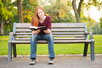 A casual young woman sitting on a bench in a park and reading her Bible.